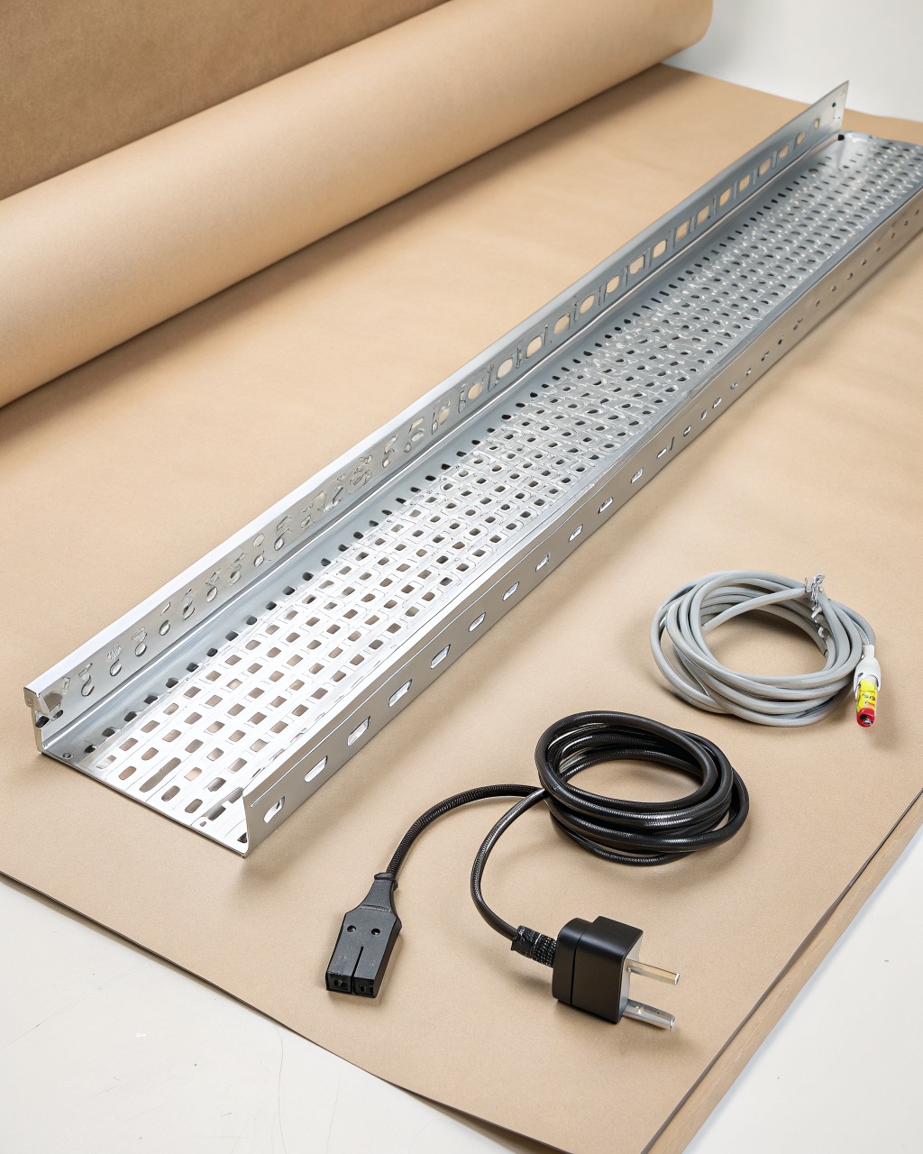 Perforated anodized-silver aluminum cable tray clamped under a desk edge holding a power strip and coiled cables