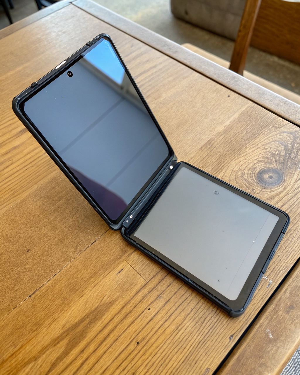 Graphite book-style foldable smartphone shown half-open, visible center crease on the large inner screen, outer cover display, triple rear camera, no branding