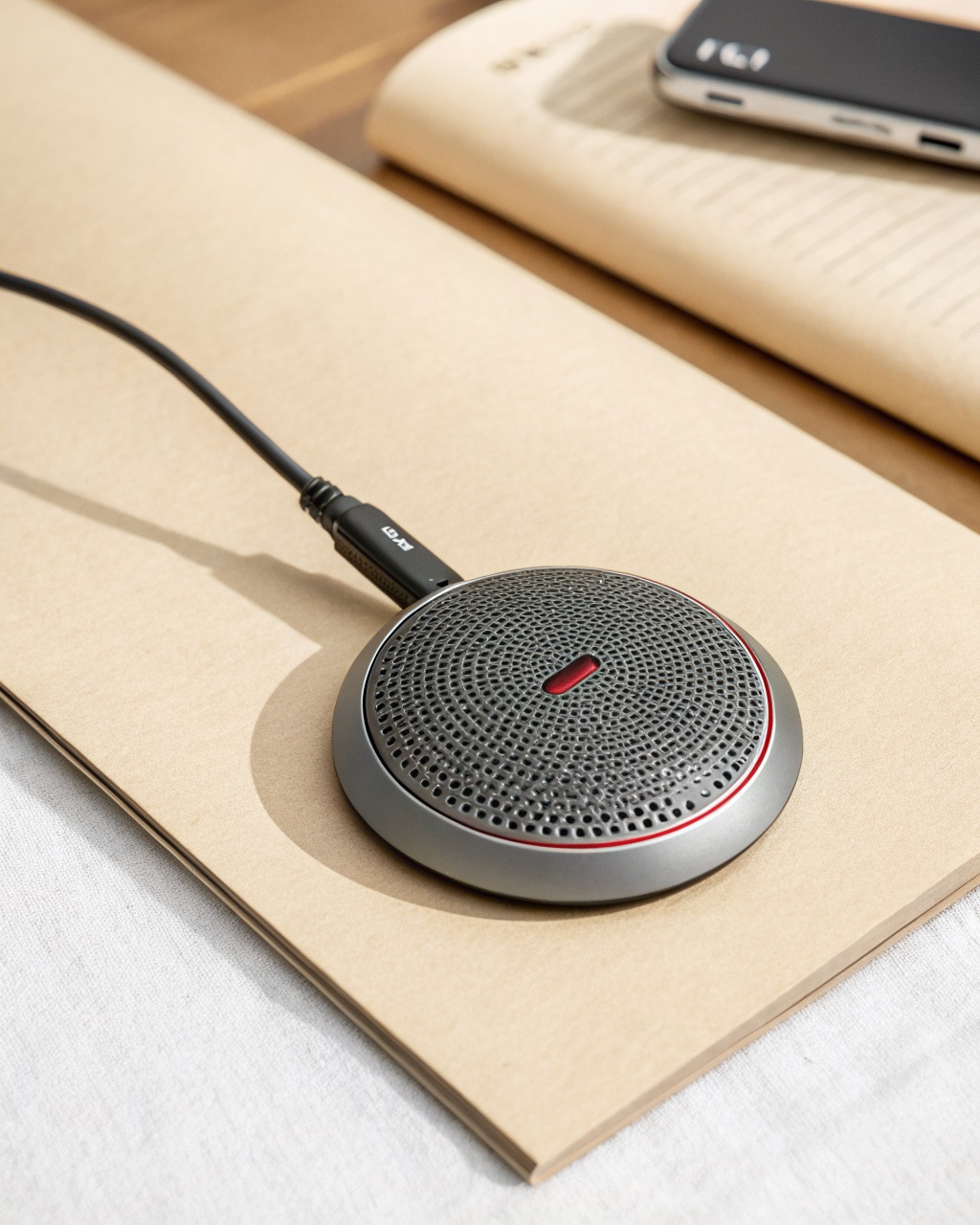 Flat disc-shaped grey USB conference microphone with a perforated metal top, a glowing touch mute ring, and a USB-C cable