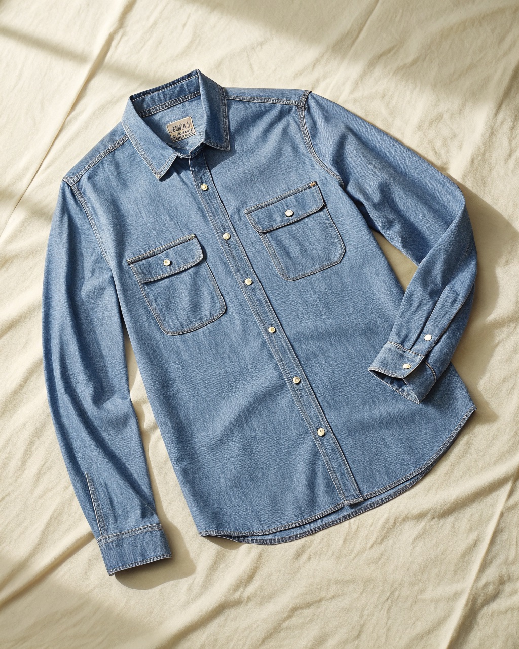 Light indigo cotton chambray work shirt laid flat with a button-down collar and two flap chest pockets