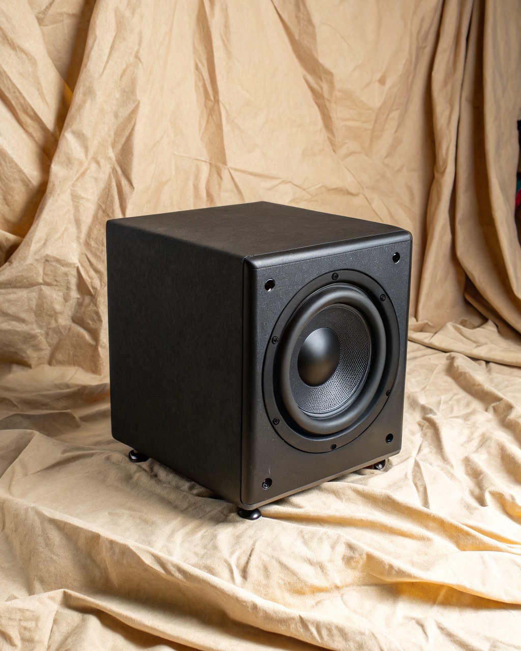Cube-shaped black ash vinyl-wrapped subwoofer with a down-firing driver, sitting on short feet on a wood floor