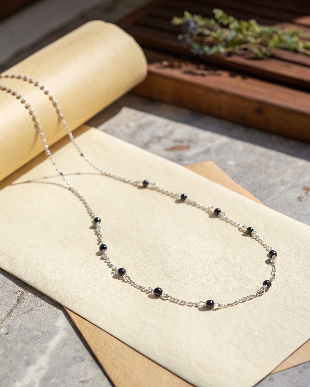 fine silver chain with small matte black onyx beads spaced along it, arranged on a pale stone surface with soft shadow