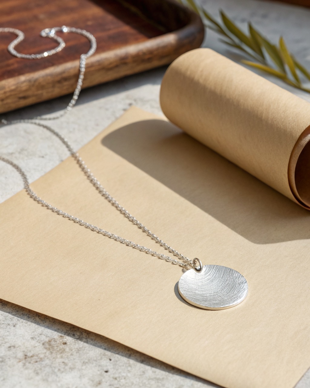 brushed sterling silver disc pendant on a fine chain lying flat on a pale stone surface, soft reflection across the disc