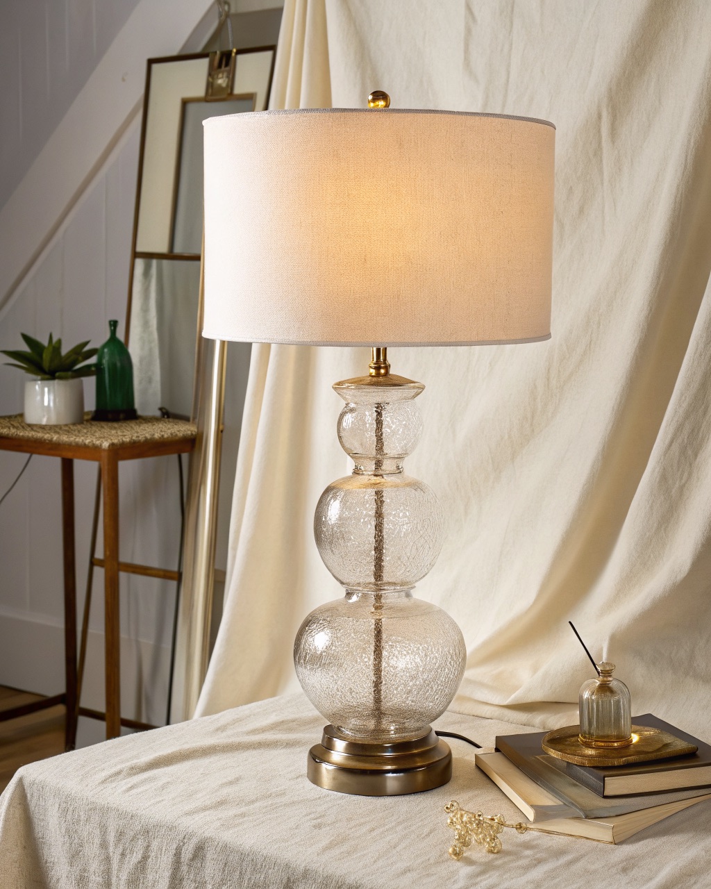 Table lamp with a hollow turned clear-glass body lit softly from within, topped by a white linen drum shade