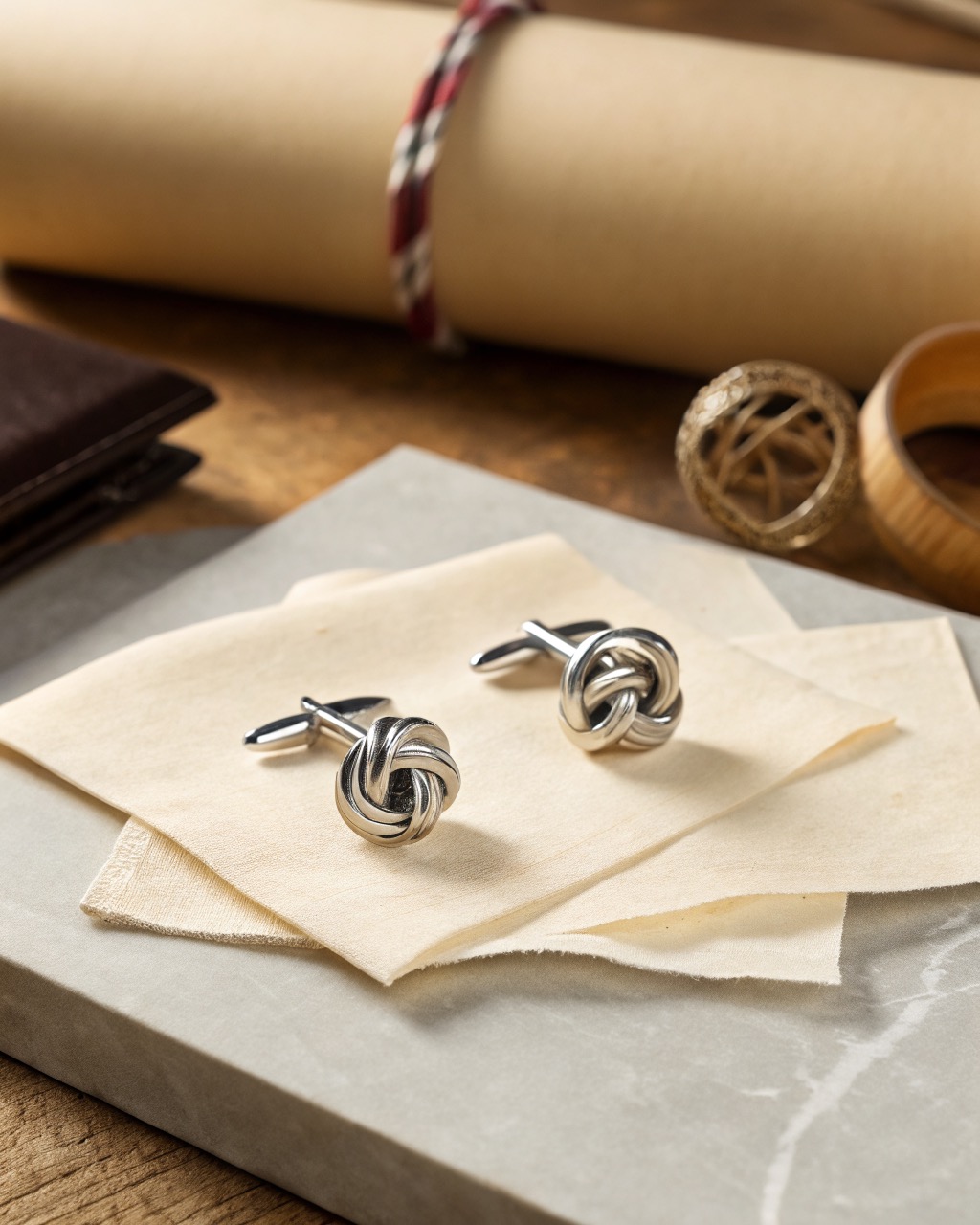 pair of polished stainless steel knot cufflinks on a pale stone surface, woven faces reflecting bright soft light