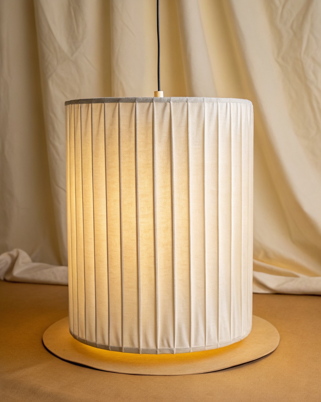 Cylindrical pendant lantern in pleated off-white linen with vertical folds and a frosted disc at the base